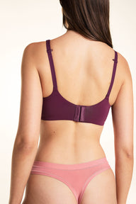 On a light studio backdrop, Laura wears the Base Unlined Bra in Eggplant. The back view showcases the smooth band, adjustable straps, and hook-and-eye closure.