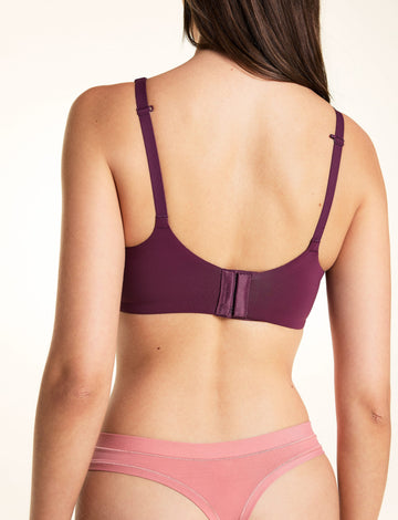 On a light studio backdrop, Laura wears the Base Unlined Bra in Eggplant. The back view showcases the smooth band, adjustable straps, and hook-and-eye closure.