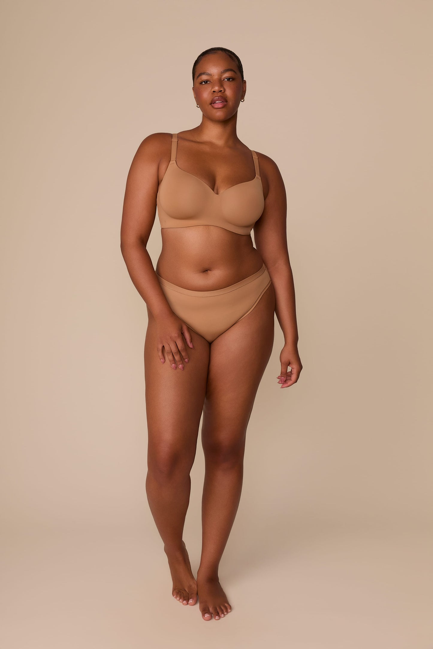 On a neutral beige backdrop, Zarria wears the Base Wirefree T-Shirt Bra in Tan with matching underwear, highlighting the smooth cups, wireless support, and clean silhouette in a full-body view.