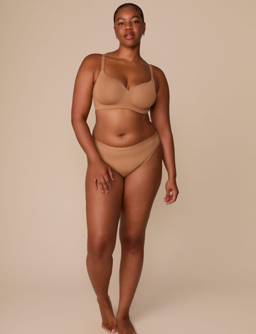 On a neutral beige backdrop, Zarria wears the Base Wirefree T-Shirt Bra in Tan with matching underwear, highlighting the smooth cups, wireless support, and clean silhouette in a full-body view.