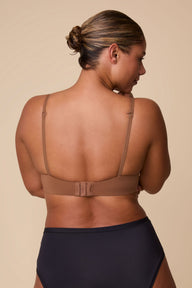 On a neutral beige backdrop, India wears the Base T-Shirt Bra in Brown, shown from the back to highlight the smooth band, adjustable straps, and hook-and-eye closure.