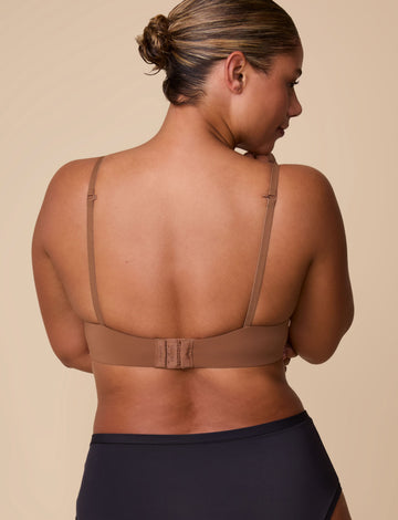 On a neutral beige backdrop, India wears the Base T-Shirt Bra in Brown, shown from the back to highlight the smooth band, adjustable straps, and hook-and-eye closure.