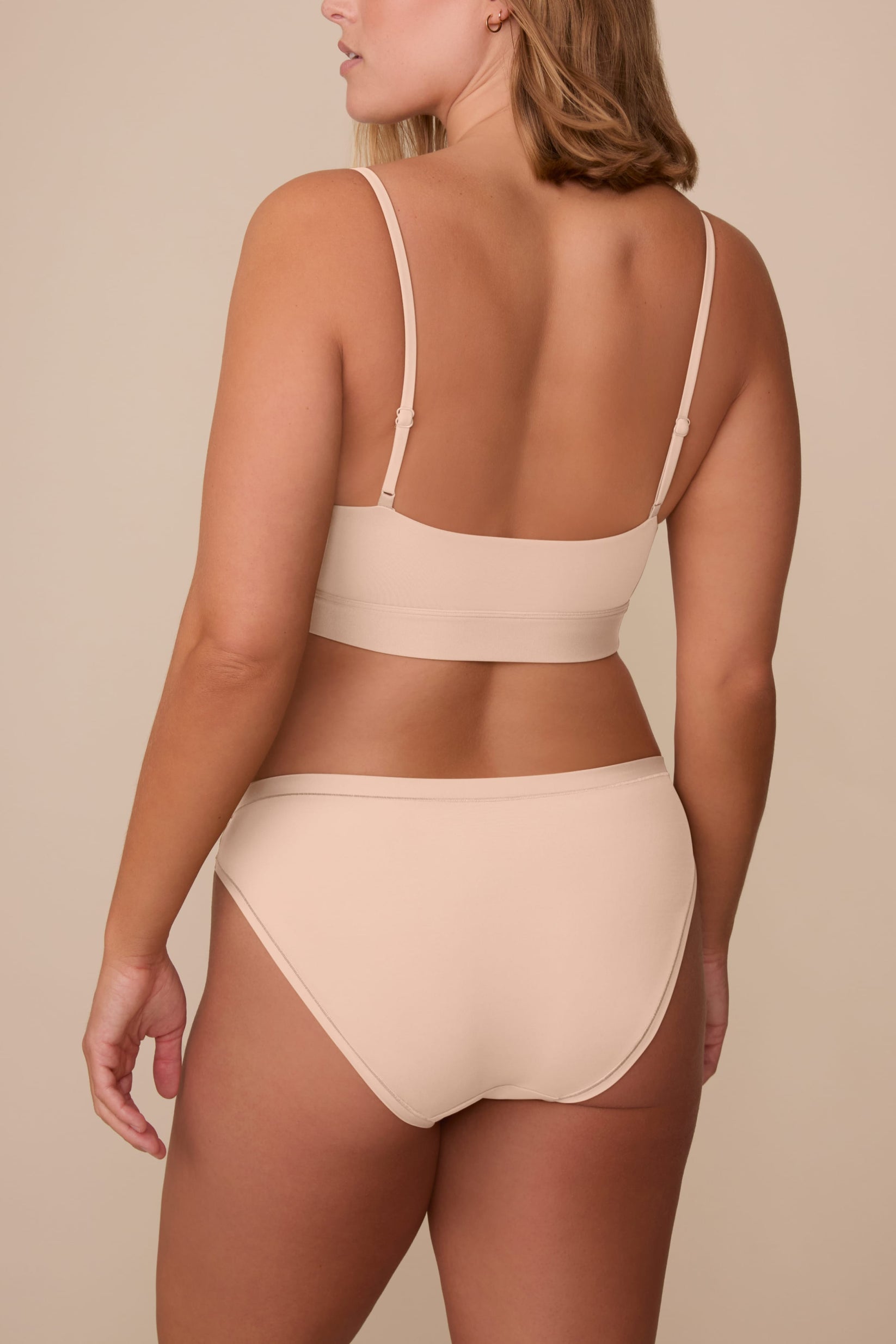 On a neutral beige backdrop, Sonnie wears the Bliss Bikini in Beige, shown from the back to highlight the full back coverage and smooth, clean finish.