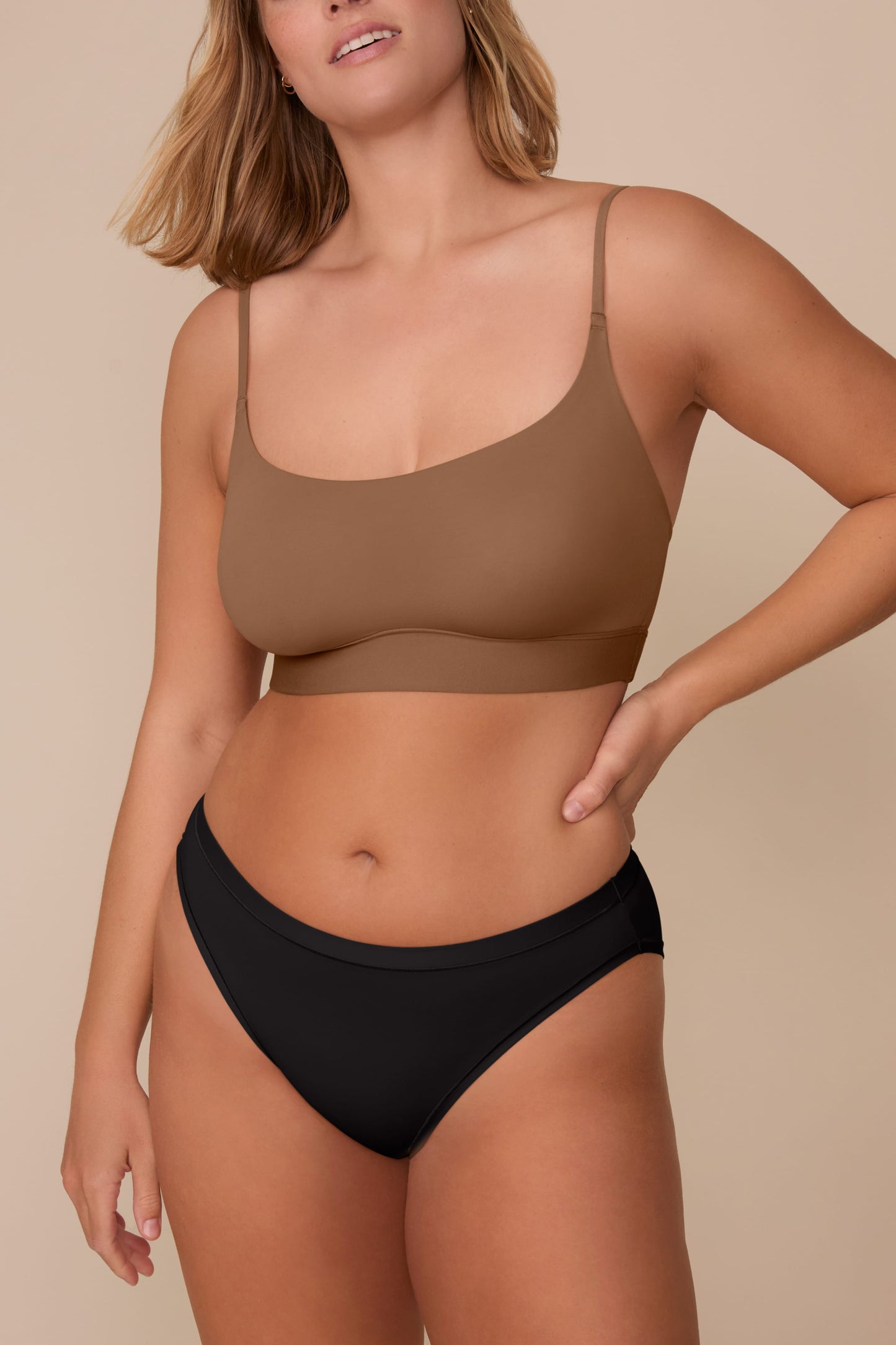 On a neutral beige backdrop, Sonnie wears the Bliss Bikini in Black with a matching bralette in a warm brown shade, showcasing the low-rise fit and smooth, minimal design.