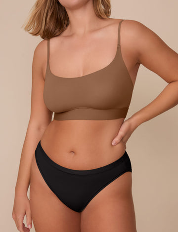 On a neutral beige backdrop, Sonnie wears the Bliss Bikini in Black with a matching bralette in a warm brown shade, showcasing the low-rise fit and smooth, minimal design.