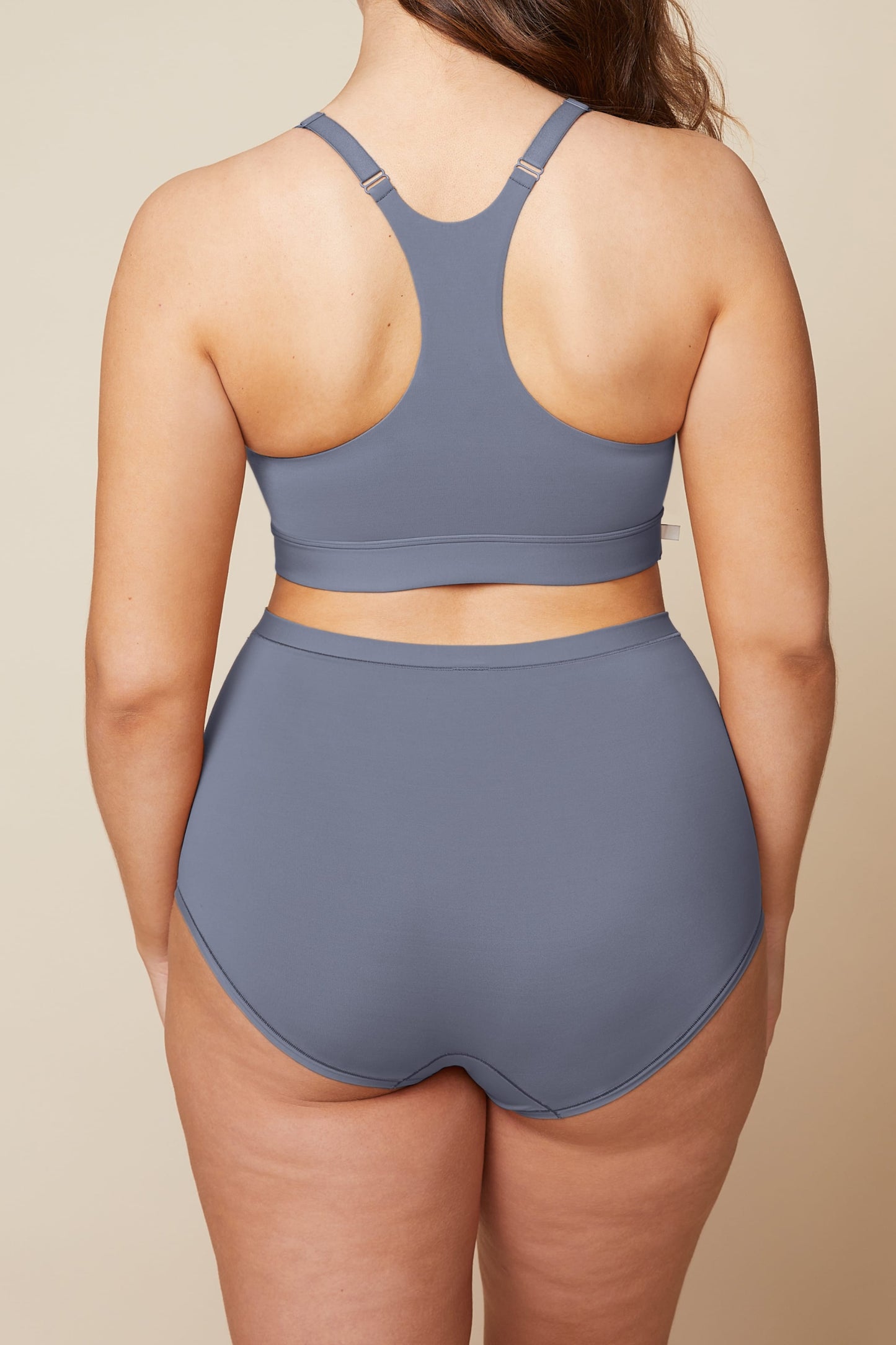 On a neutral beige backdrop, a back view of Summerlyn wearing the Bliss Boyshort in Steel highlights the full-coverage back and smooth, high-rise waistband.