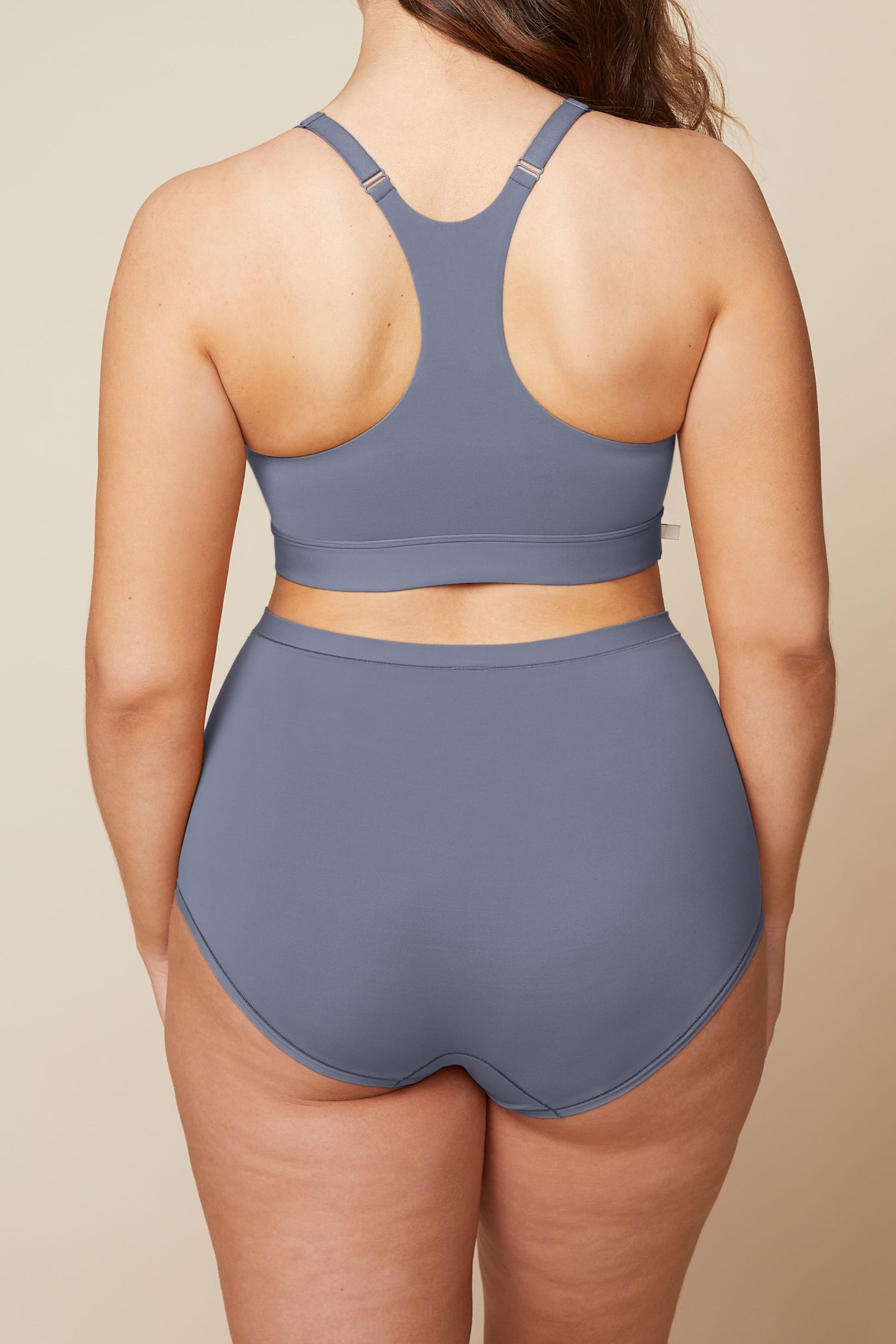 On a neutral beige backdrop, a back view of Summerlyn wearing the Bliss Boyshort in Steel highlights the full-coverage back and smooth, high-rise waistband.