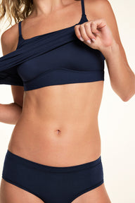 On a light studio backdrop, Alexis lifts the Bliss Cami in Navy to reveal the built-in bra. The close-up highlights the scoop neckline, supportive underbust band, and smooth fabric construction.