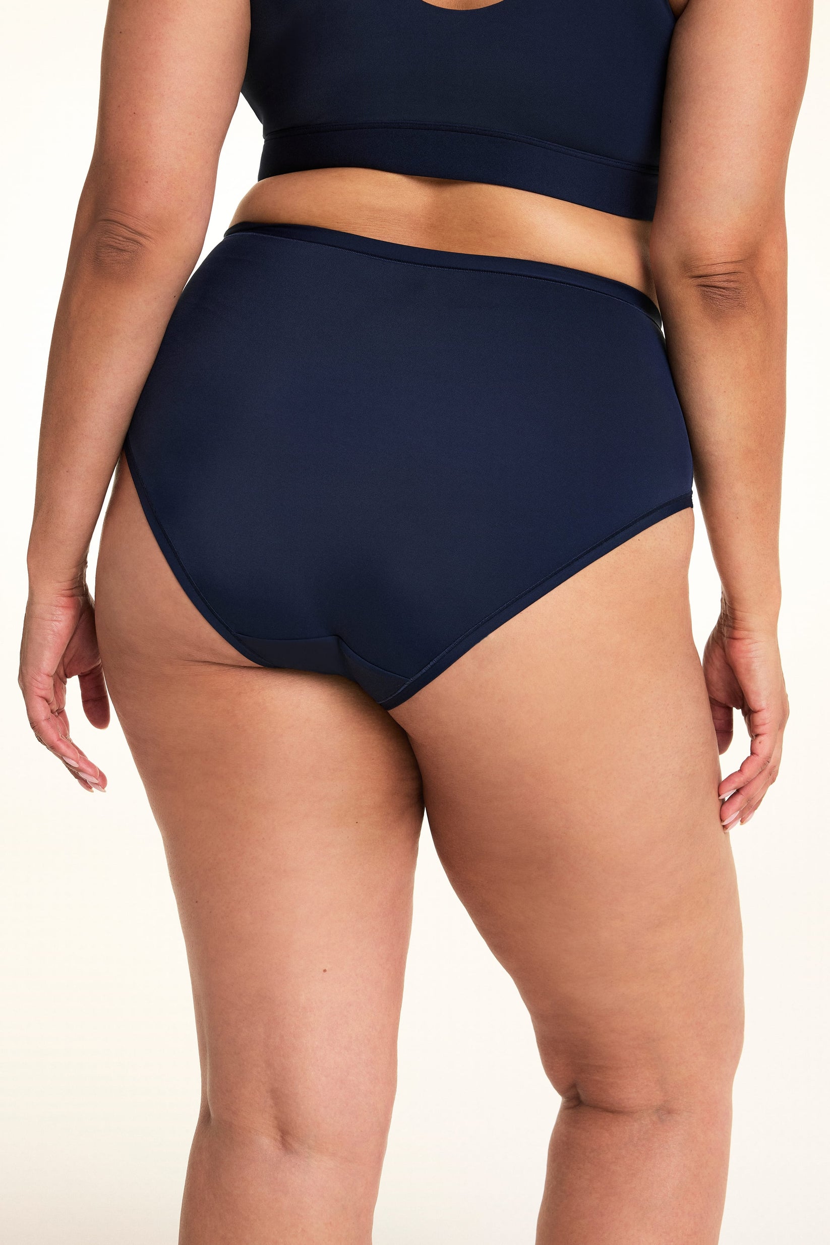 On a light studio backdrop, Jennifer is shown from the back wearing the Bliss Highwaist in Navy. The close-up highlights the full-coverage back, smooth fabric, and clean leg openings.