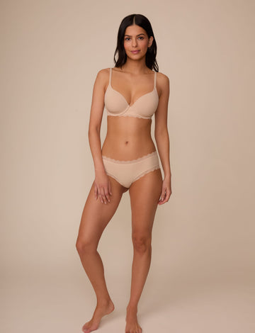 On a neutral studio backdrop, Marcela wears the Bliss Lace Cheeky in Beige with a matching beige bra. The full-body front view showcases the smooth fabric, low-rise fit, and delicate scalloped lace detailing at the waist and leg openings.