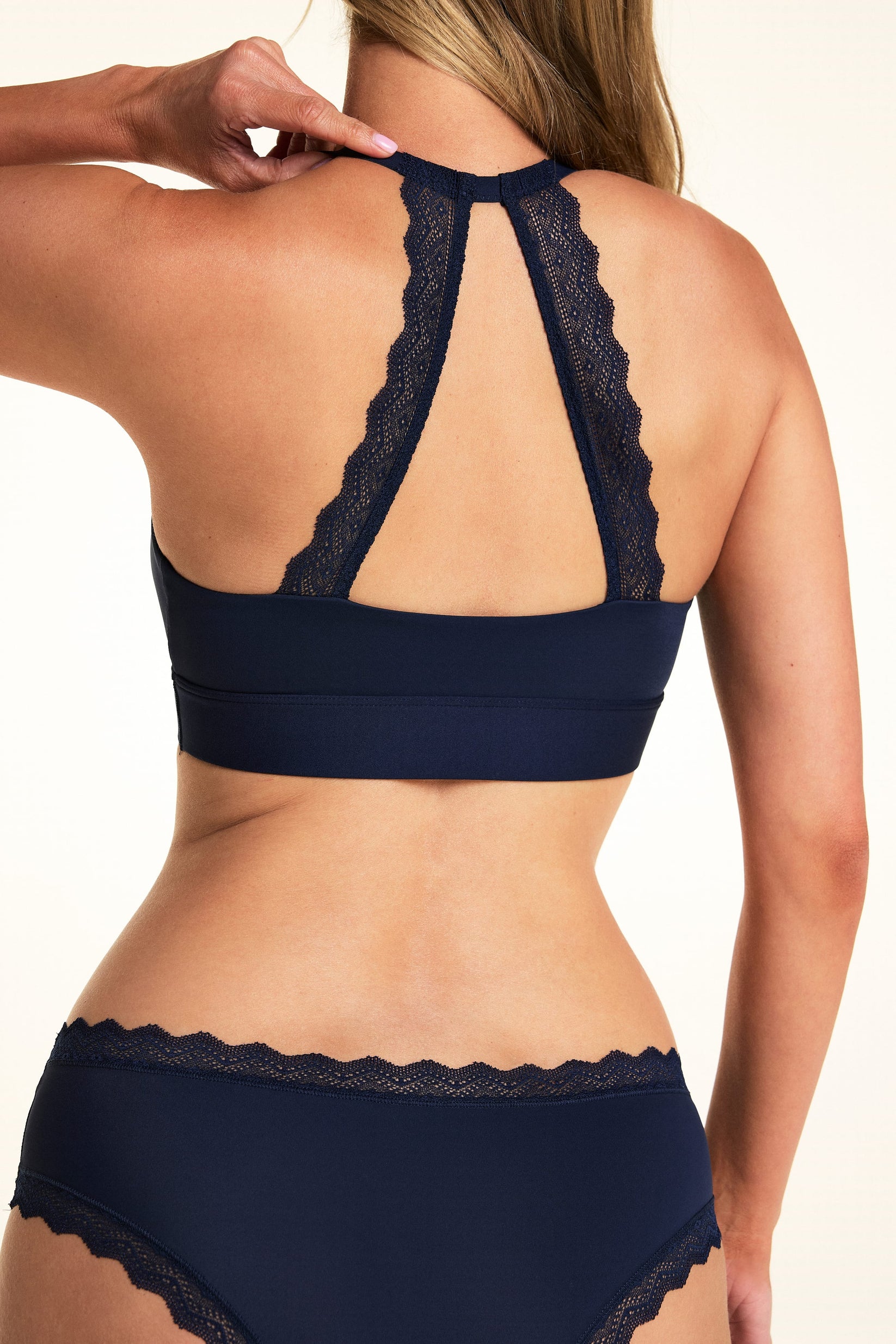On a light studio backdrop, Meagan is shown from the back wearing the Bliss Lace Halterneck Bralette in Navy. The lace halter back detail creates an open keyhole design, showcasing the scalloped lace trim and wide underband.