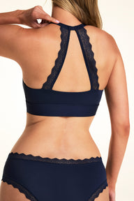On a light studio backdrop, Meagan is shown from the back wearing the Bliss Lace Halterneck Bralette in Navy. The lace halter back detail creates an open keyhole design, showcasing the scalloped lace trim and wide underband.