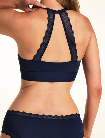 On a light studio backdrop, Meagan is shown from the back wearing the Bliss Lace Halterneck Bralette in Navy. The lace halter back detail creates an open keyhole design, showcasing the scalloped lace trim and wide underband.