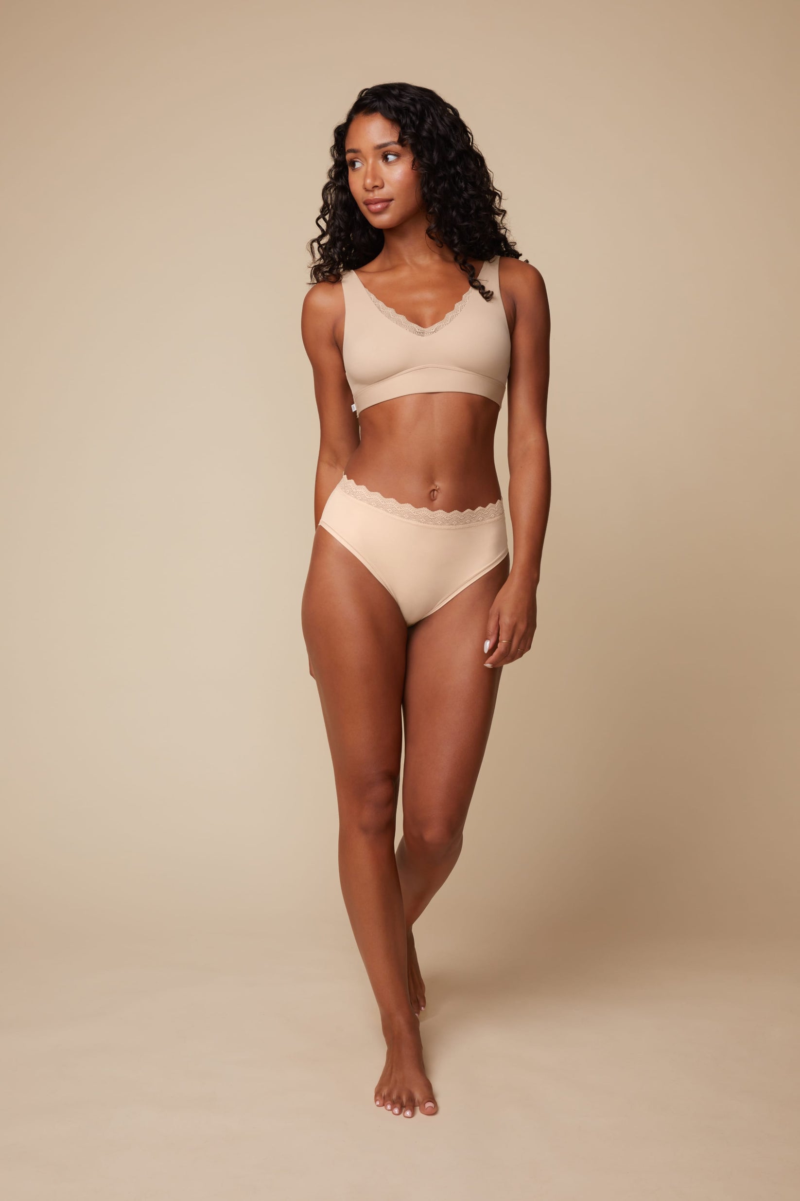 On a neutral beige backdrop, Ally wears the Bliss Lace Highwaist Brief in Beige with the matching Bliss Lace Bralette, highlighting the smooth fabric, high-waisted fit, and lace trim at the waist.