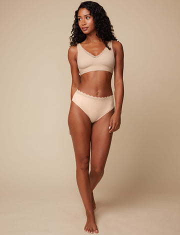 On a neutral beige backdrop, Ally wears the Bliss Lace Highwaist Brief in Beige with the matching Bliss Lace Bralette, highlighting the smooth fabric, high-waisted fit, and lace trim at the waist.
