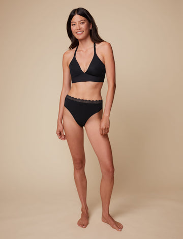 On a neutral beige backdrop, Anna wears the Bliss Lace Highwaist Thong in Black with the matching Bliss Lace Bralette, highlighting the high-waisted fit and lace-trim waistband in a full-body view.