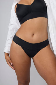 In a studio setting, India wears the Bliss Low Rise Thong in Black with the matching Bliss Bralette in Black. A close-up front view highlights the smooth waistband, low-rise fit, and minimal-coverage thong design.