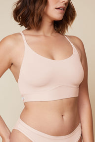 On a neutral beige backdrop, a close-up of Carli wearing the Bliss Racerback Bralette in Rose highlights the smooth fabric, scoop neckline, adjustable straps, and supportive underband.