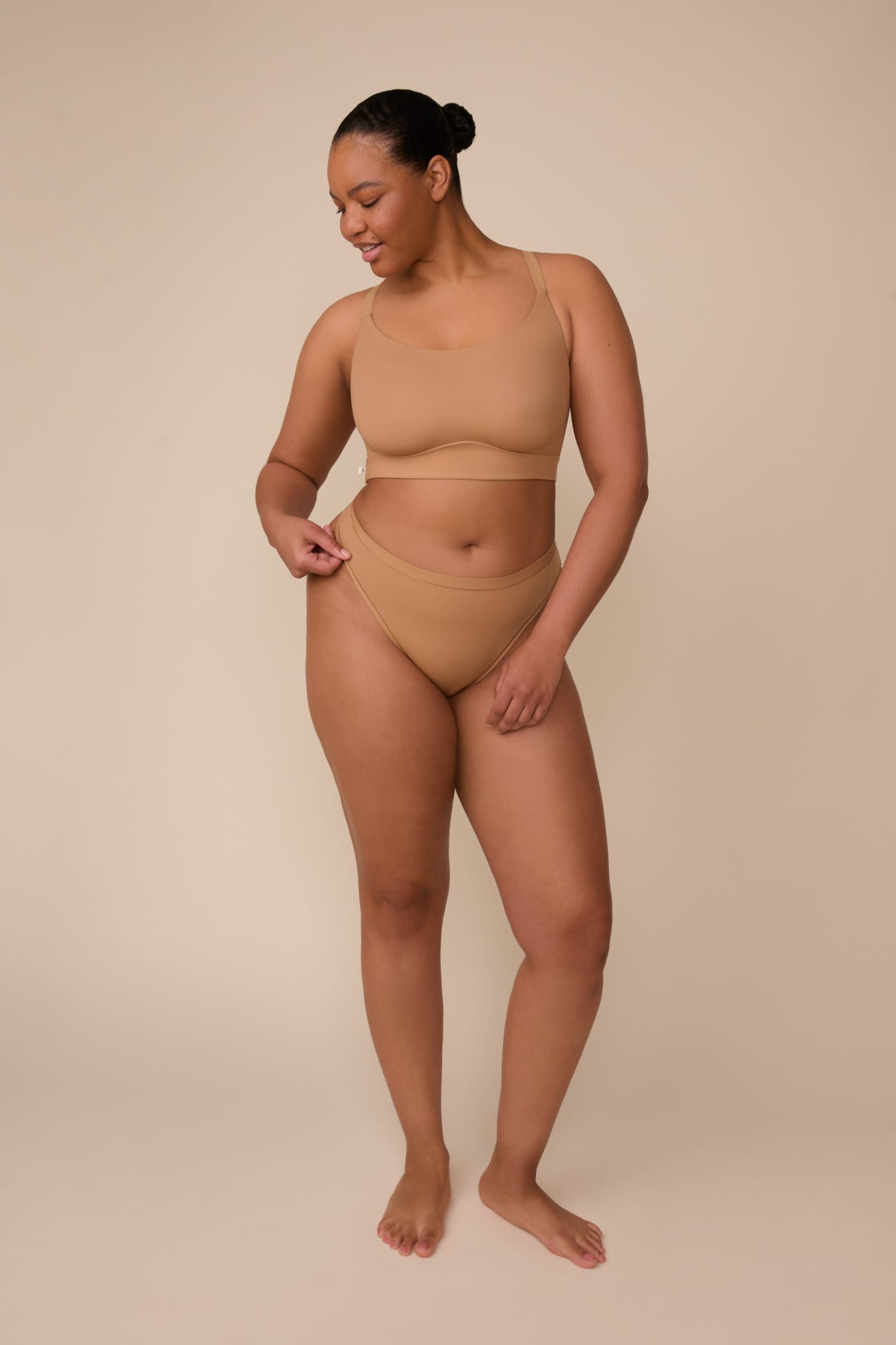 On a neutral beige backdrop, Zarria wears the Bliss Racerback Bralette in Tan with coordinating underwear, showcasing the scoop neckline, adjustable straps, and smooth fit while gently holding the waistband.
