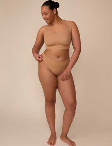 On a neutral beige backdrop, Zarria wears the Bliss Racerback Bralette in Tan with coordinating underwear, showcasing the scoop neckline, adjustable straps, and smooth fit while gently holding the waistband.