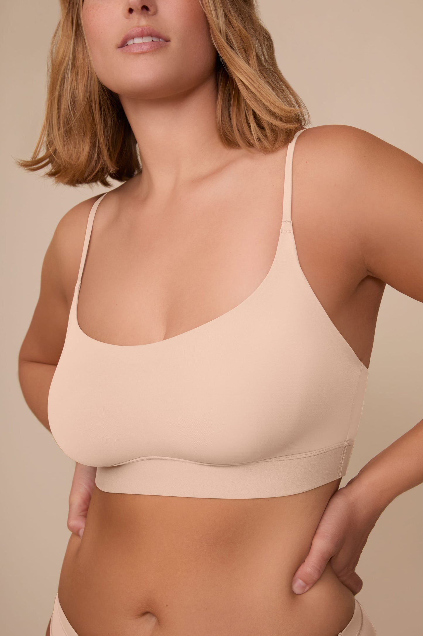 On a neutral beige backdrop, a close-up of Sonnie wearing the Bliss Scoop Adjustable Bralette in Beige showcases the scoop neckline, adjustable straps, and smooth fabric for a comfortable fit.