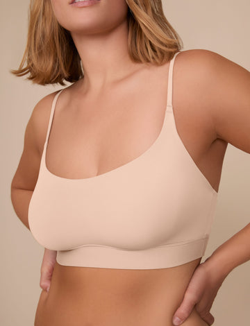On a neutral beige backdrop, a close-up of Sonnie wearing the Bliss Scoop Adjustable Bralette in Beige showcases the scoop neckline, adjustable straps, and smooth fabric for a comfortable fit.