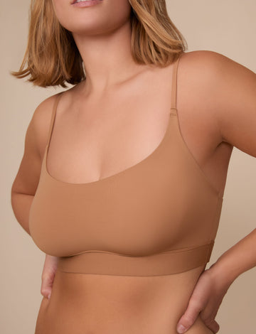 On a neutral beige backdrop, a close-up of Sonnie wearing the Bliss Scoop Adjustable Bralette in Tan showcases the scoop neckline, adjustable straps, and smooth fabric for a comfortable fit.