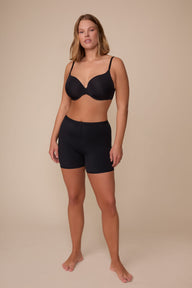 On a neutral studio backdrop, Sonnie wears the Bliss Under Short in Black with a matching black bra. The full-body front view showcases the smooth fabric, mid-rise fit, and lightweight thigh coverage.
