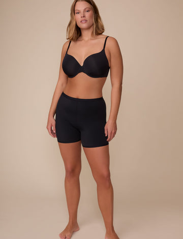 On a neutral studio backdrop, Sonnie wears the Bliss Under Short in Black with a matching black bra. The full-body front view showcases the smooth fabric, mid-rise fit, and lightweight thigh coverage.