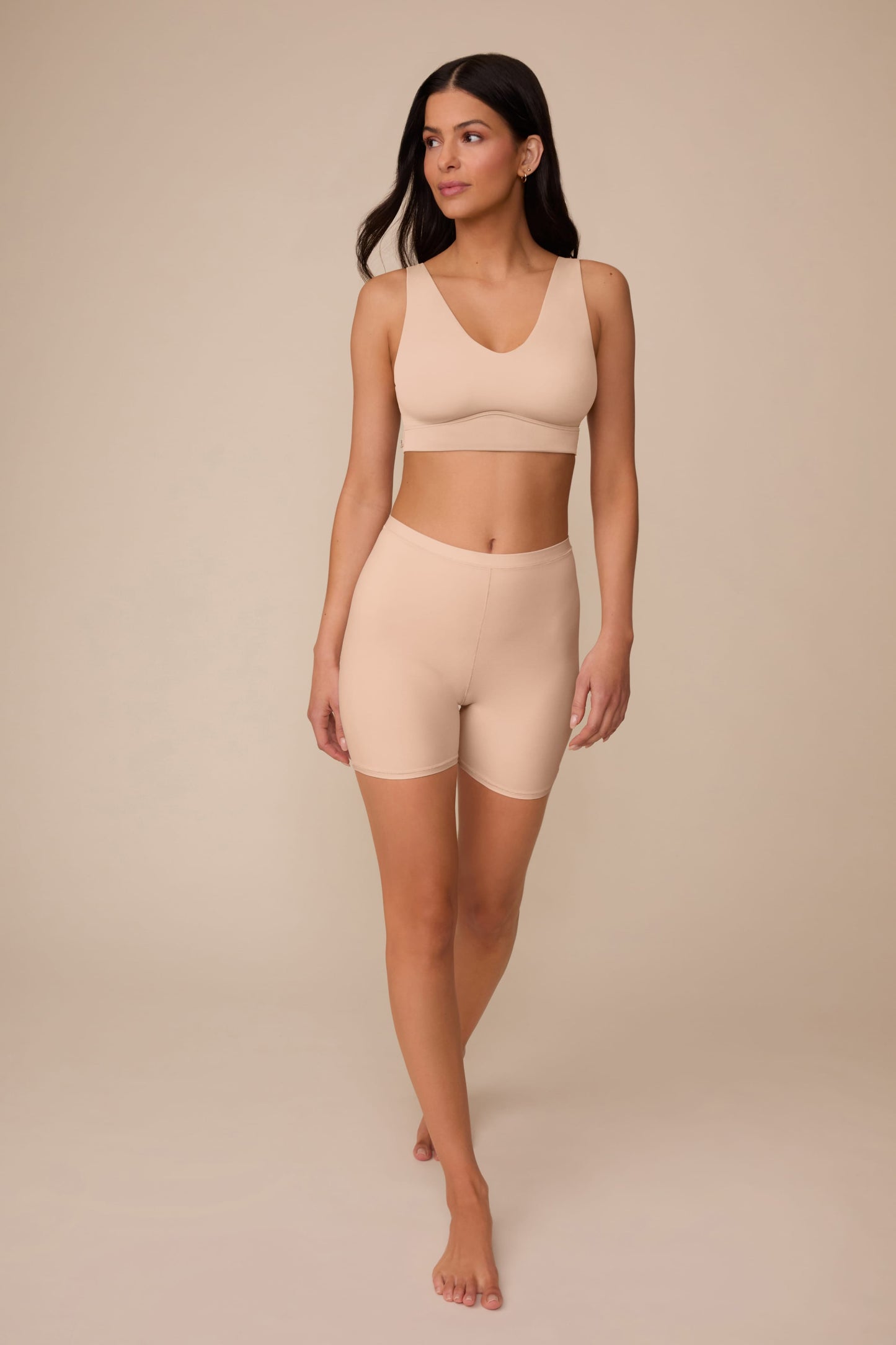 On a neutral studio backdrop, Marcela wears the Bliss Under Short in Beige with a matching beige bralette. The full-body view showcases the lightweight fabric, smooth finish, and thigh coverage.