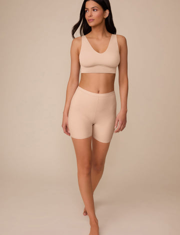 On a neutral studio backdrop, Marcela wears the Bliss Under Short in Beige with a matching beige bralette. The full-body view showcases the lightweight fabric, smooth finish, and thigh coverage.