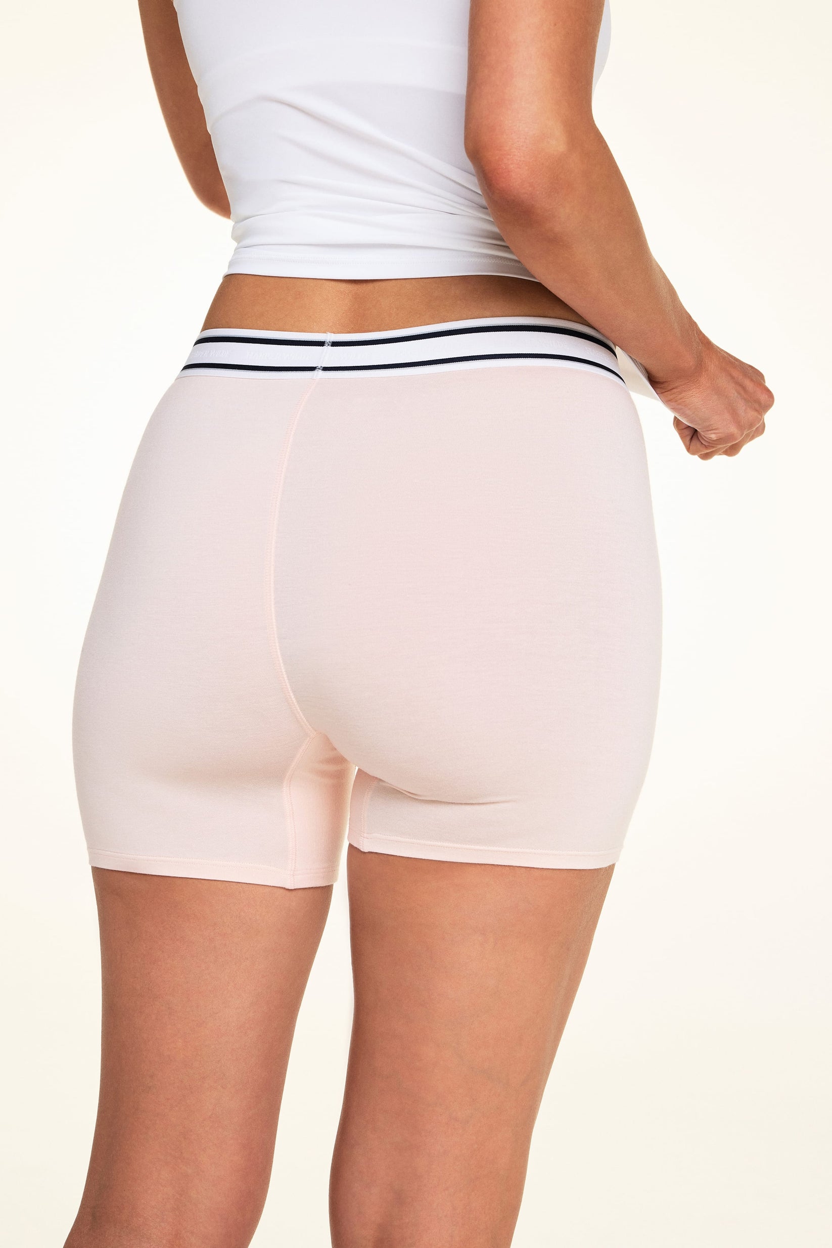 On a light studio backdrop, Meagan wears the Cloud Cotton Boxer Brief in Dogwood with a white tank top. The back view highlights the full-coverage boxer brief silhouette, smooth fabric, and striped elastic waistband.