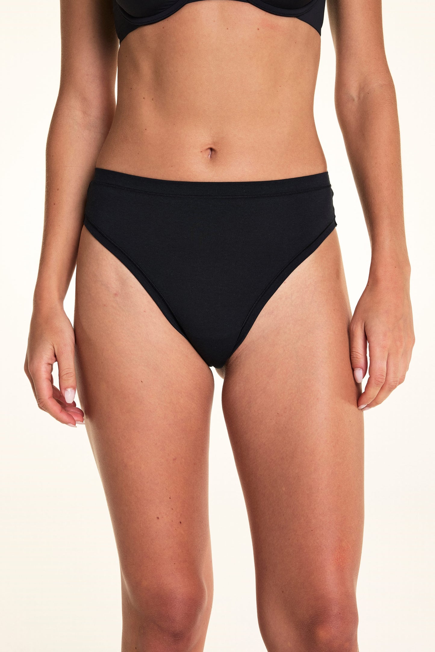Against a white studio backdrop, Alexis wears the Cloud Cotton Highwaist Thong in Black, shown from the front to highlight the high-rise waistband and soft cotton design.
