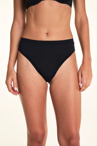 Against a white studio backdrop, Alexis wears the Cloud Cotton Highwaist Thong in Black, shown from the front to highlight the high-rise waistband and soft cotton design.