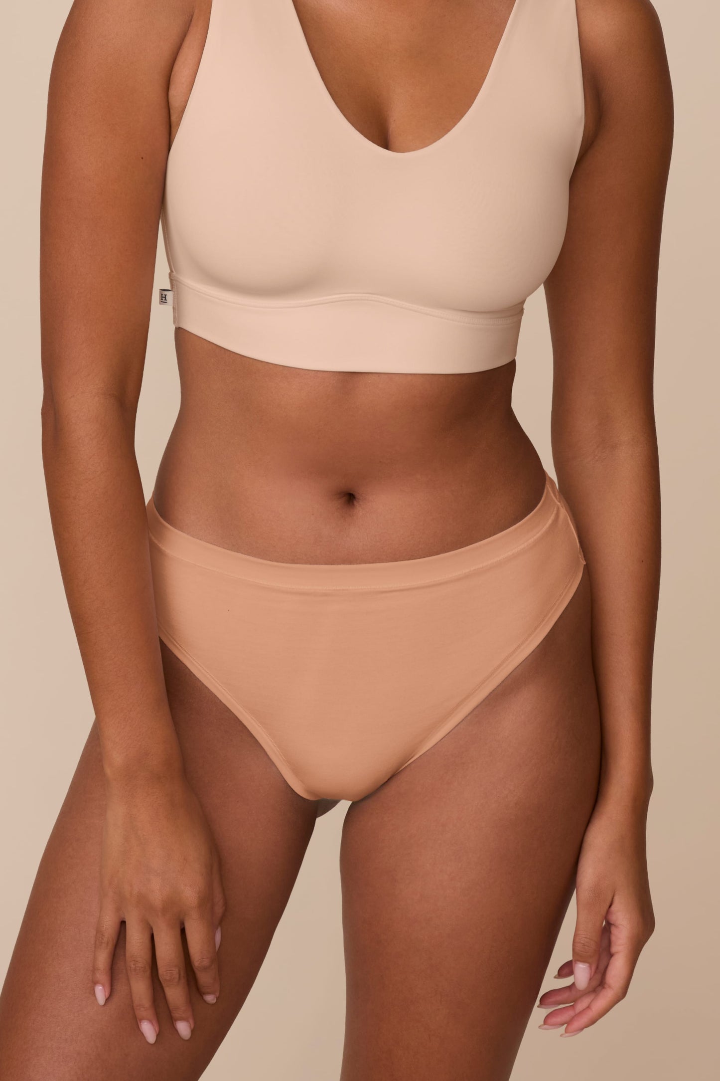 On a neutral beige backdrop, a close-up of Victoria wearing the Cloud Cotton Highwaist Thong in Dune highlights the high-rise waistband and soft cotton fabric.
