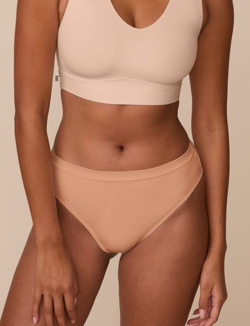 On a neutral beige backdrop, a close-up of Victoria wearing the Cloud Cotton Highwaist Thong in Dune highlights the high-rise waistband and soft cotton fabric.