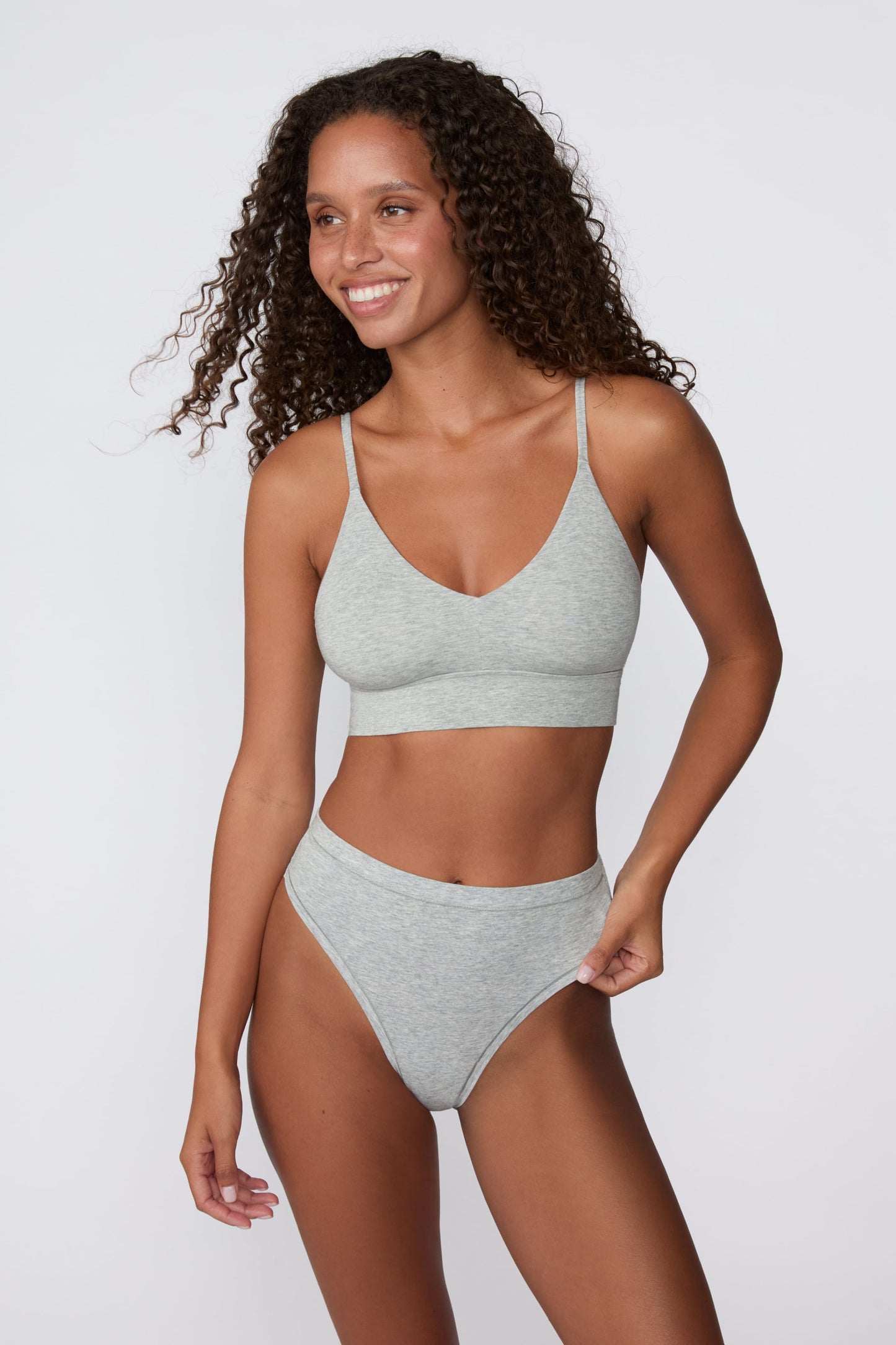On a light gray studio backdrop, Courtney wears the Cloud Cotton Highwaist Thong in Heather Grey with the matching bralette. The front view highlights the high-rise waistband, smooth fit, and high-leg silhouette.