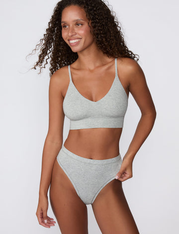 On a light gray studio backdrop, Courtney wears the Cloud Cotton Highwaist Thong in Heather Grey with the matching bralette. The front view highlights the high-rise waistband, smooth fit, and high-leg silhouette.