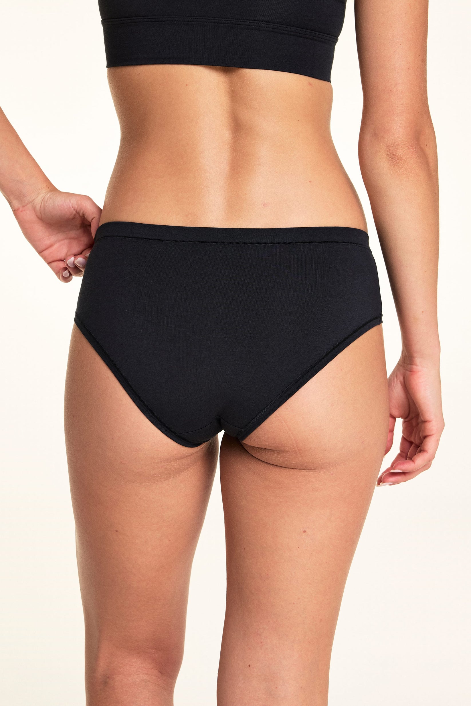 Captured from the back, Alexis wears the Cloud Cotton Hiphugger underwear in Black while gently pulling the waistband to show the flexible fit and smooth back 