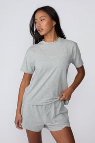 On a light gray studio backdrop, Amber wears the Cloud Cotton Pajama Shirt in Heather Grey with matching pajama shorts. The front three-quarter view showcases the relaxed fit, crew neckline, short sleeves, and lightweight cotton fabric.
