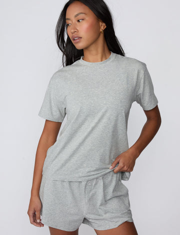On a light gray studio backdrop, Amber wears the Cloud Cotton Pajama Shirt in Heather Grey with matching pajama shorts. The front three-quarter view showcases the relaxed fit, crew neckline, short sleeves, and lightweight cotton fabric.