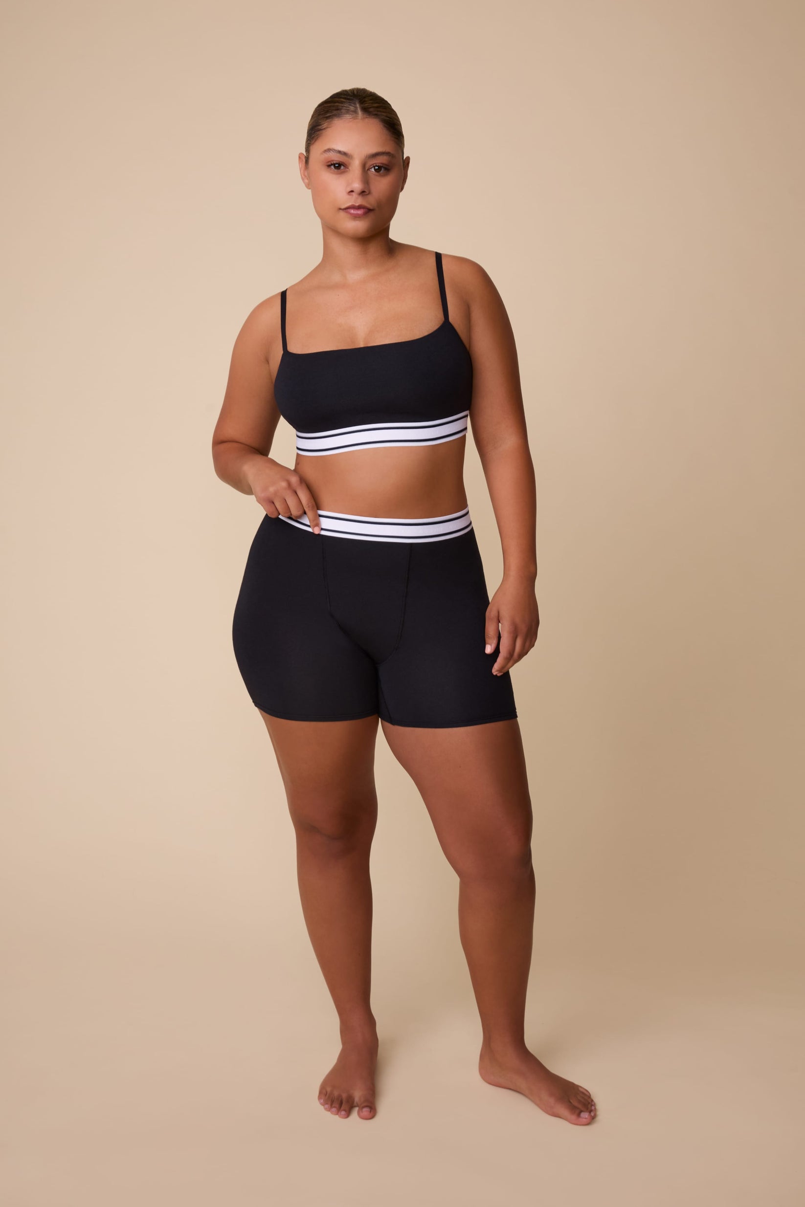 On a neutral beige backdrop, India wears the Cloud Cotton Retro Bandeau Bralette in Black with matching shorts, showcasing the straight neckline, slim adjustable straps, and striped elastic waistband details.