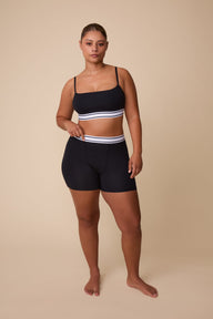 On a neutral beige backdrop, India wears the Cloud Cotton Retro Bandeau Bralette in Black with matching shorts, showcasing the straight neckline, slim adjustable straps, and striped elastic waistband details.
