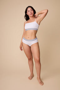 On a neutral beige backdrop, Emersyn wears the Cloud Cotton Retro Bandeau Bralette in White with matching underwear, highlighting the smooth bandeau silhouette, adjustable straps, and striped elastic underband in a full-body view. sizing: Emersyn wears size Large