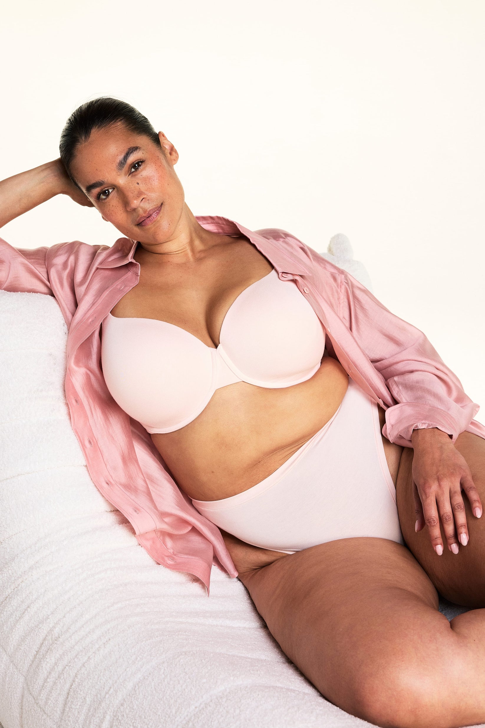 In a cozy bedroom setting, Jennifer lounges on a soft white chair wearing the Cloud Cotton T-Shirt Bra in Dogwood paired with matching underwear and an open pink button-down shirt. The image highlights the smooth molded cups, underwire support, and comfortable everyday design.