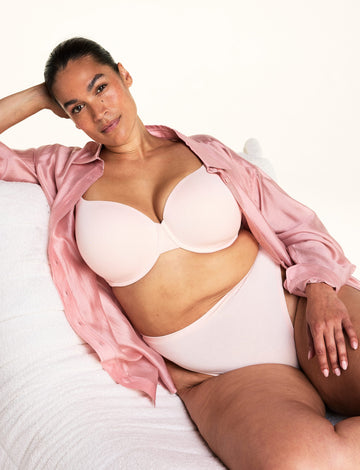In a cozy bedroom setting, Jennifer lounges on a soft white chair wearing the Cloud Cotton T-Shirt Bra in Dogwood paired with matching underwear and an open pink button-down shirt. The image highlights the smooth molded cups, underwire support, and comfortable everyday design.