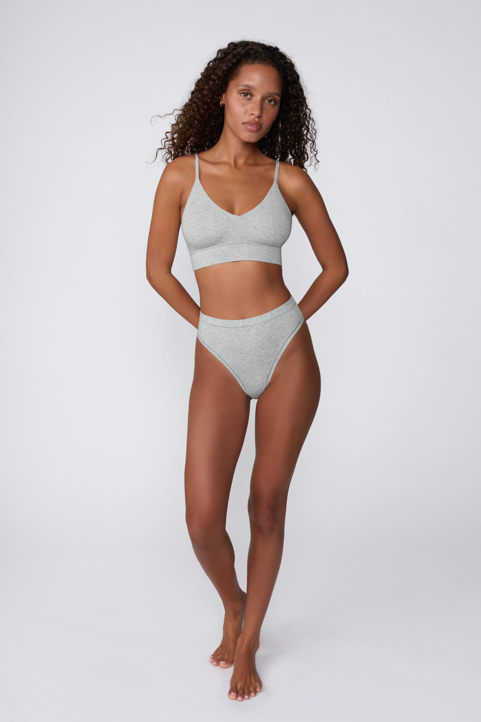 In a studio setting, Courtney wears the Cloud Cotton Triangle Bralette in Heather Grey with matching underwear. The full-body view highlights the soft cotton fabric, deep V-neckline, and wide underband.