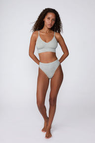 In a studio setting, Courtney wears the Cloud Cotton Triangle Bralette in Heather Grey with matching underwear. The full-body view highlights the soft cotton fabric, deep V-neckline, and wide underband.