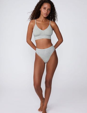 In a studio setting, Courtney wears the Cloud Cotton Triangle Bralette in Heather Grey with matching underwear. The full-body view highlights the soft cotton fabric, deep V-neckline, and wide underband.