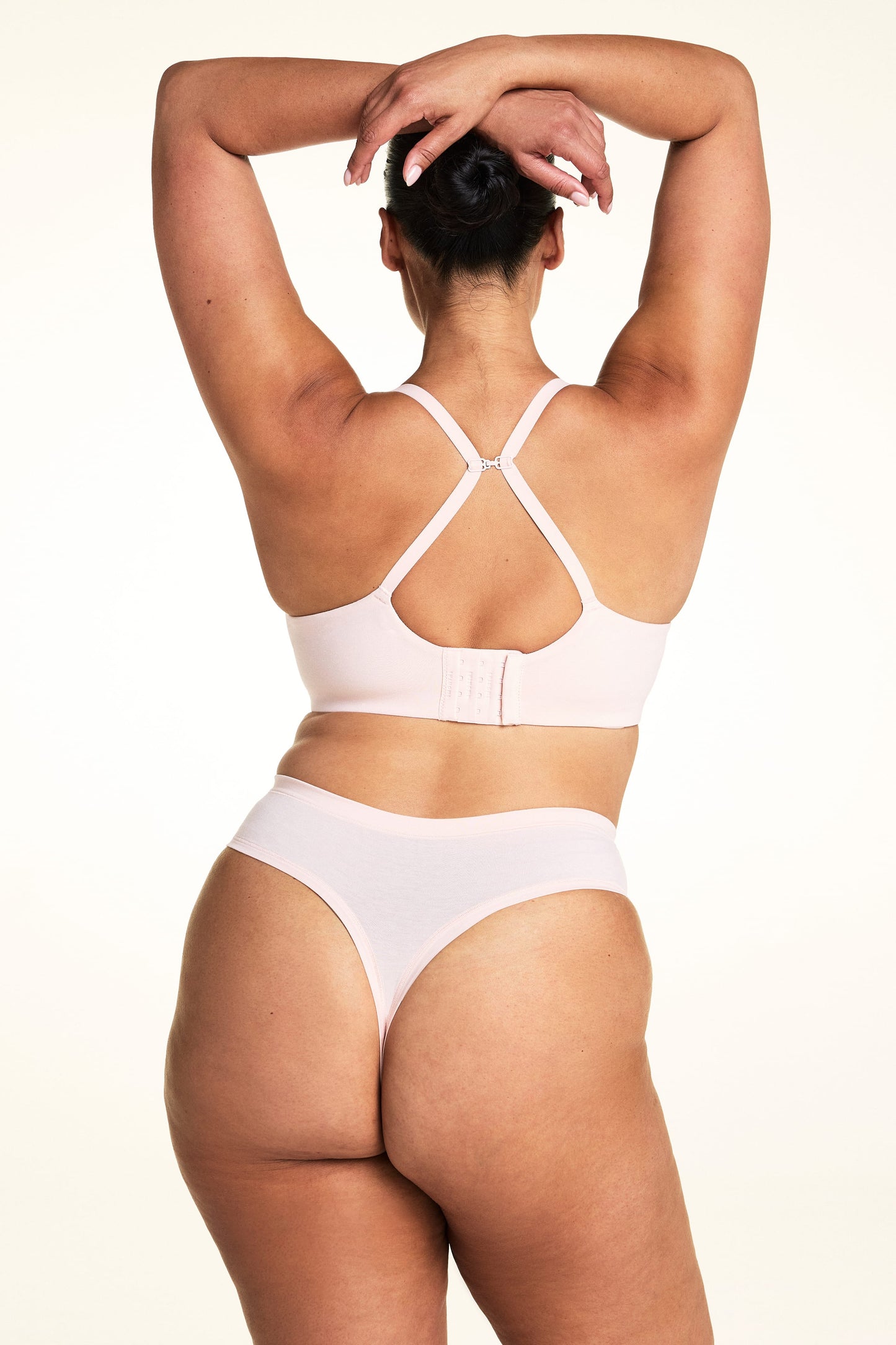 On a light studio backdrop, Jennifer wears the Cloud Cotton Highwaist Thong in Dogwood with a matching bra in Dogwood. The back view highlights the thong back coverage, smooth waistband, and convertible racerback straps on the bra.